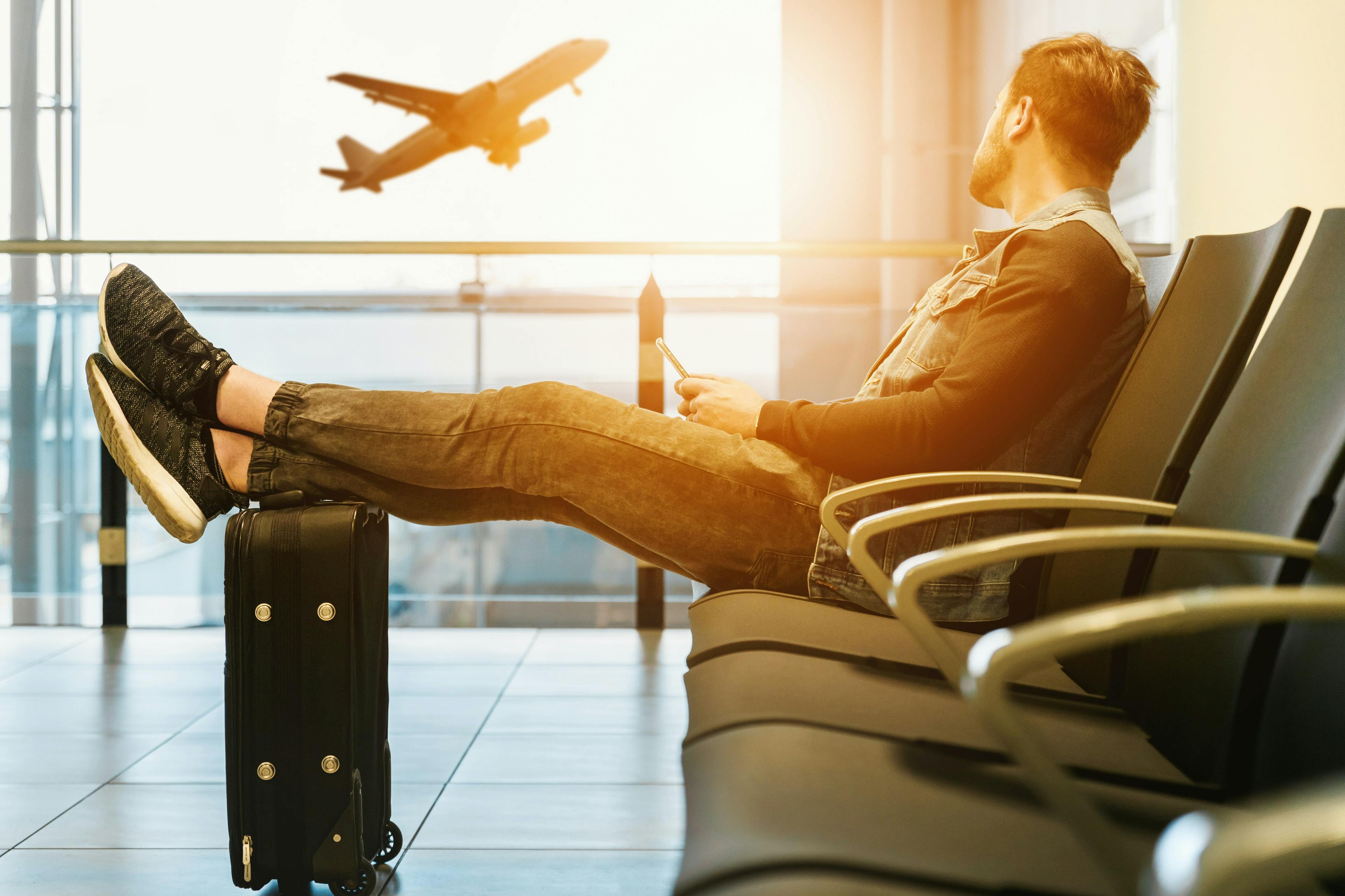 a man is seating in a chair in an airport lounge, resting his legs in his luggage while looking through the window at an airplane that's taking off